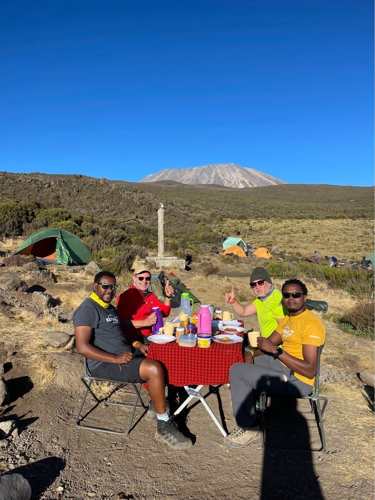 Group of climbers enjoying a meal at a campsite on Mount Kilimanjaro with tents and the mountain peak in the background